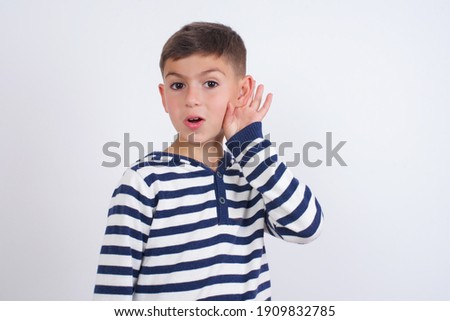 Oh my God! Funny astonished little cute Caucasian boy kid wearing stripped t-shirt against white wall opening mouth widely looking aside, with hand near ear trying to listen to gossips. Royalty-Free Stock Photo #1909832785