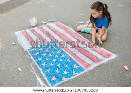 Girl drawing American flag with colored chalks on the sidewalk near the house on sunny summer day. Kids painting outside. Creative development of children.