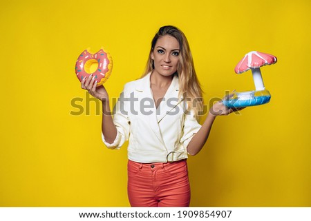 Attractive fit woman with a great figure and blond hair holding small inflatable beach toys in her hands. Flapper toys umbrella and blue and pink donuts. On a yellow isolated background. 