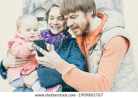Happy parents showing picture on phone to toddler girl. Family with baby walking near old construction outdoors. Communication concept