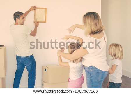 Caucasian dad in casual clothes hanging up empty frame on wall in new home and smiling. Blonde mother and two daughters helping him with aligning picture. Family, relocation and moving day concept