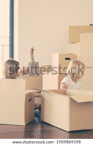 Two little girls unpacking things in new apartment, sitting on floor and taking objects from open cartoon boxes. Relocation or moving concept