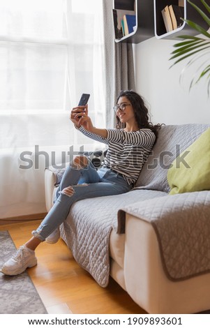 Young girl with glasses sitting in her living room, she is taking a selfie with her phone.