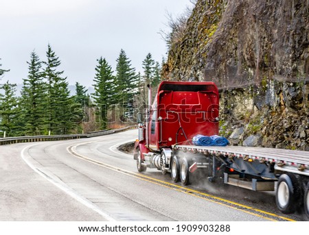 Big rig red semi truck with chrome pipes transporting empty flat bed semi trailer running on the winding wet road with rain dust driving on the bridge with rock cliff on the side in Columbia Gorge