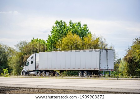 Big rig white long haul industrial semi truck tractor with sleeper cab compartment for truck driver rest transporting cargo in dry van semi trailer driving on the highway road with summer trees
