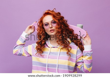 Beautiful redhead woman in lilac glasses looks into camera and holds skateboard on isolated background