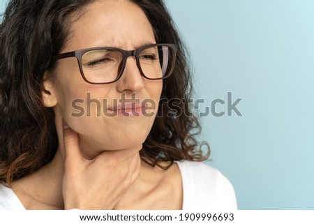 Closeup of sick woman wear glasses having sore throat, tonsillitis, caught cold, suffering from painful swallowing, strong pain in throat, holding hand on her neck, isolated on studio blue background Royalty-Free Stock Photo #1909996693