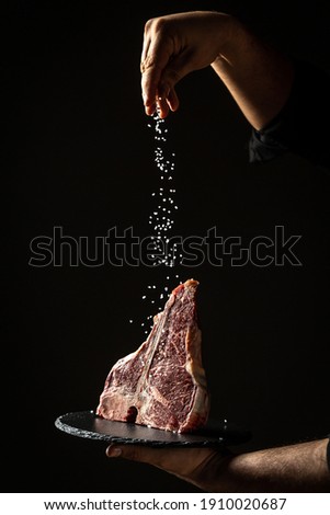 Chef hands cooking dry aged wagyu porterhouse beef steak with large fillet piece adding salt and pepper in a freeze motion on black background. vertical image, place for text.