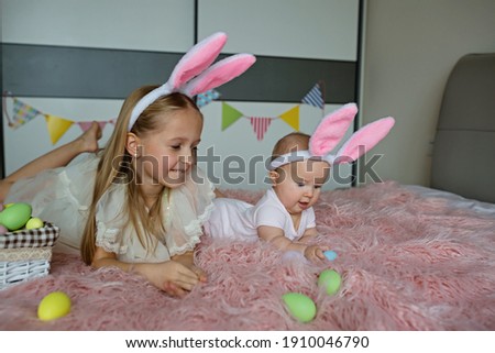 happy caucasian toddler girl 7 years old and her baby sister 6 months old wearing bunny ears headband, lying on bed at home in bedroom with colored easter eggs. Stay home during Coronavirus covid-19