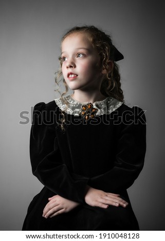 Beautiful young girl in black 1890s English Victorian 18th century child period dress with elegant white lace collar antique broach jewelery and long curly pretty hair looking away from camera Royalty-Free Stock Photo #1910048128