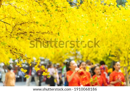 Decorative plastic yellow apricot flowers in Tet holiday in Vietnam. People wear Vietnam tradition ao dai to take pictures in blurred background. Background and festival concept. Selective focus. 