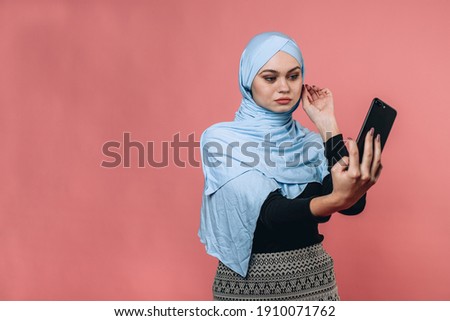Cute young girl in a black jacket and patterned skirt and dressed in a light blue hijab holding a black iPhone in her right hand posing for a photo. Search for angle for a story on vlog. Blog concept 