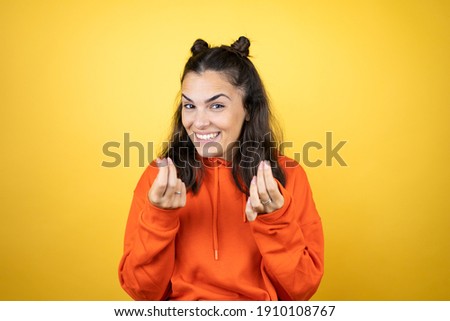 Young beautiful woman wearing sweatshirt over isolated yellow background doing money gesture with hands, asking for salary payment, millionaire business