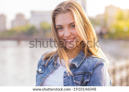 Portrait of a happy and attractive blonde Caucasian young woman in a casual denim jacket outdoors in a park on a sunny autumn day. A concept of lifestyle, happiness and joy. Royalty-Free Stock Photo #1910146048