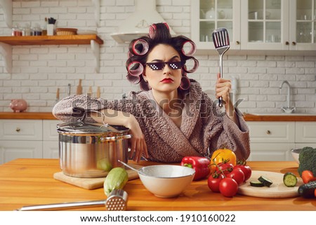 Funny angry young housewife in bad mood cooking lunch and waiting for husband at home. Woman in bathrobe, curlers, thug life 8 bit pixel sunglasses holding spatula sitting at kitchen table like a boss Royalty-Free Stock Photo #1910160022
