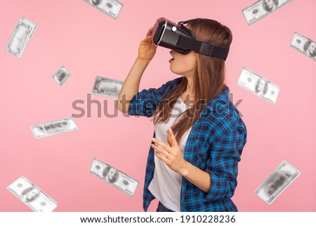 Profile side view portrait of shocked woman wearing virtual reality glasses, looking at money fall, dollar rain, illusion of rich millionaire, playing vr game, indoor isolated on pink background