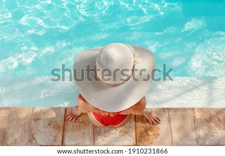 Top view of young woman in red one-piece swimsuit and straw hat relax near a swimming pool with legs in water
