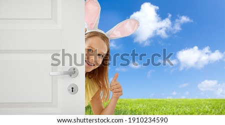 easter, holidays and people concept - happy girl wearing bunny ears peeking out door and showing thumbs up over blue sky and grass background