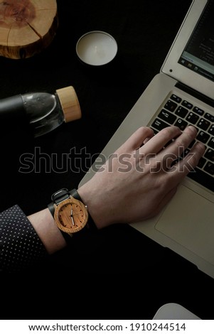 A stylish wooden watch on the hand of a young man who is typing on a laptop surrounded by a bottle of water, a candle and a wooden stump. Picture in a low key.