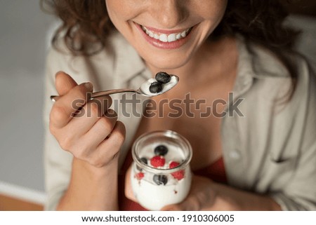 Close up of of smiling woman having a healthy breakfast at home with fruit and yogurt. Girl tasting yoghurt with blueberries and raspberries. Woman enjoy fresh yogurt for lunch, wellbeing diet concept Royalty-Free Stock Photo #1910306005