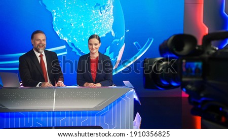 Live News Studio with Beautiful Female and Handsome Man Anchors Start Reporting. TV Broadcasting Channel with Presenters Talking. Television Newsroom Set. Behind the Scene Camera Shooting Shot Royalty-Free Stock Photo #1910356825