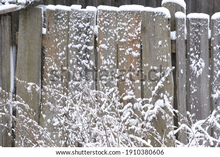 Wooden fence gate covered in white snow at heavy snowing snowstorm, storm, falling snowflakes, bushes in background. Snow on a wooden fence as background image. No edit.