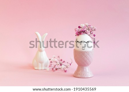 Easter egg with a black marker drawn face in a pink vase with flowers and bunny on pink .