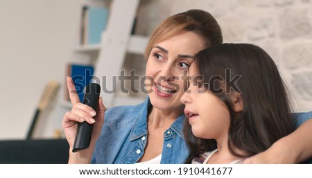 Cute little daughter and young mother are sitting on the sofa. She chooses a program on TV. Smiling mother holding remote control watching cartoons on TV with little boy at home.