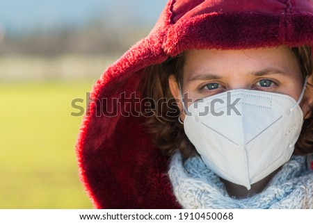 Close up portrait of young woman with blue eyes wearing red hodded jacket in a green field wearing face mask