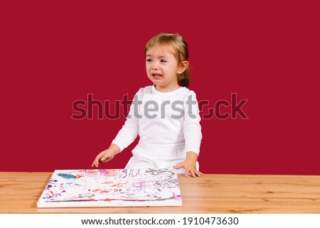 Little girl crying sitting at the wooden table with canvas picture set. Unpatient toddler girl waiting her mummy come to create painting by numbers