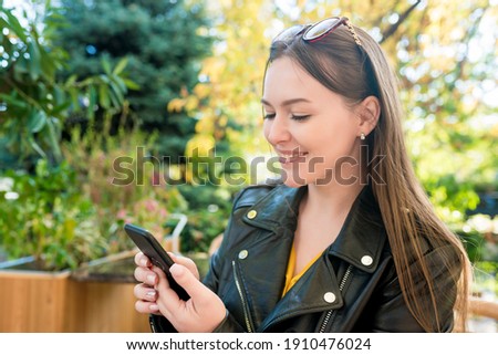  Cute young woman in casual clothes sitting in cafe outside on sunny day using smartphone. People texting on mobile during brunch at restaurant