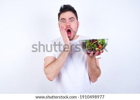 Scared terrified Young handsome Caucasian man holding a salad bowl against white background shocked with prices at shop, People and human emotions concept