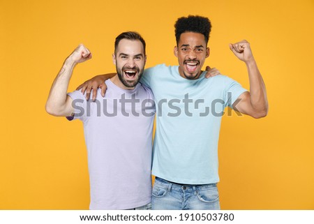 Happy joyful young friends european african american men 20s in casual violet blue t-shirts hugging doing winner gesture looking camera isolated on bright yellow colour background studio portrait