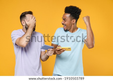 Displeased happy young two friends european african american men 20s in t-shirts play games on mobile phones cover face with palm doing winner gesture isolated on yellow background studio portrait