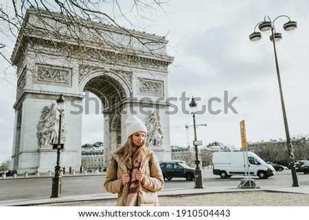 Young beautiful fashionable woman tourist in  white hat and scarf on background of  famous Arc de Triomphe or Triumphal arch. Winter or autumn in Europe. Paris, France.