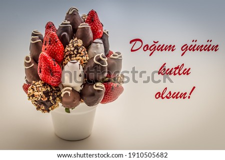 Happy birthday greeting card in Turkish Language with red lettering that reads Dogum gunun kutlu olsun; A bunch of edible flowers, arrangement of chocolate dipped strawberries isolated on white