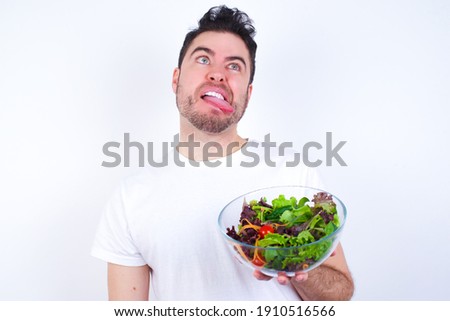 Young handsome Caucasian man holding a salad bowl against white background showing grimace face crossing eyes and showing tongue. Being funny and crazy