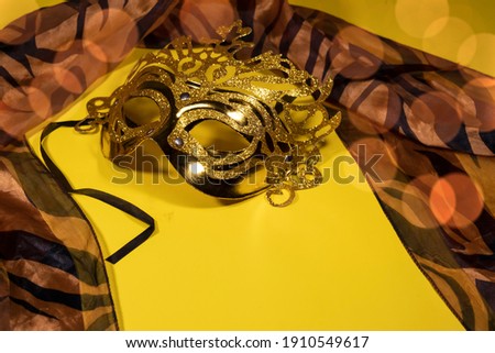 On a yellow background with bokeh, there is a silk scarf and a gilded carnival mask. 