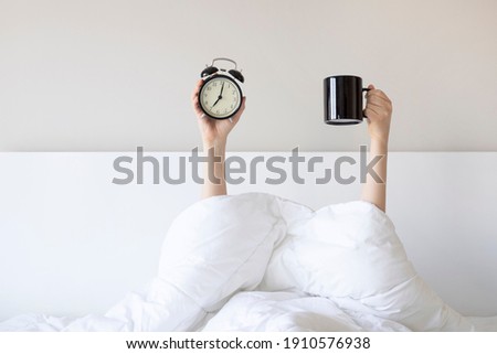 Woman showing arm raised up holding coffee cup and black alarm clock behind duvet in the bed room, Young girl with two hands sticking out from the blanket. wake up with fun in morning concept. Royalty-Free Stock Photo #1910576938