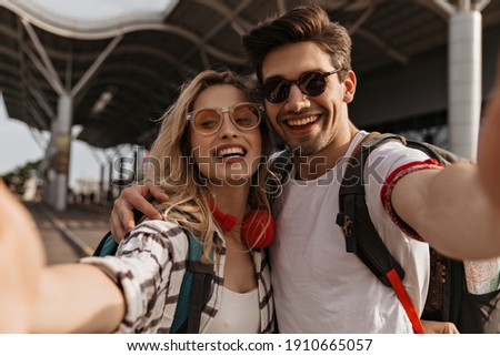 Couple if travelers takes selfie near airport. Young blonde curly woman in red headphones and brunette man in sunglasses smiles and hugs.