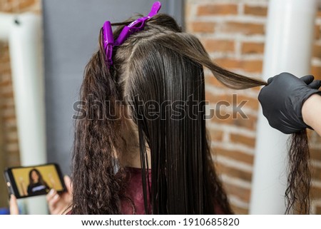 A young woman watches a video on her phone while sitting in a hair salon for a keratin hair straightening procedure. The concept of spa services and hair care. Beauty salon Royalty-Free Stock Photo #1910685820