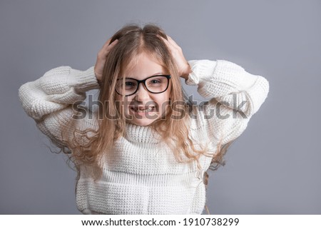 Portrait of a young attractive little girl with hands on head in a white sweater annoyed irritated pulling out her hair for nervous disagreement on a gray studio background.