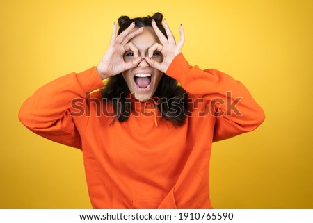 Young beautiful woman wearing sweatshirt over isolated yellow background doing ok gesture shocked with smiling face, eye looking through fingers