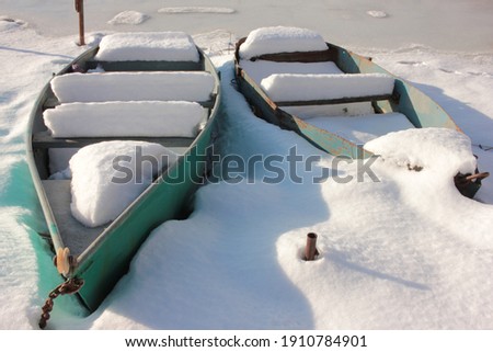 Old rusty fishing boats covered with snow on the banks of a frozen river on a sunny day. Late winter or early spring landscape. Selective focus
