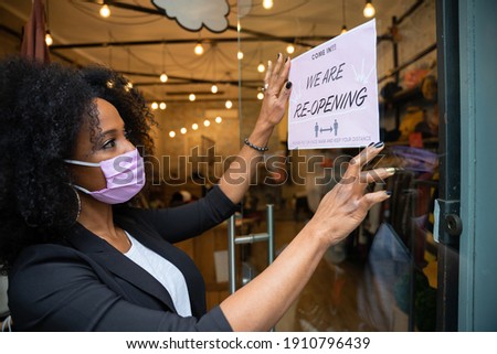 Portrait of a beautiful young owner of a clothing store while hanging a sign in the window for the reopening of the business wearing the protective face mask against Coronavirus infections, Covid-19 Royalty-Free Stock Photo #1910796439