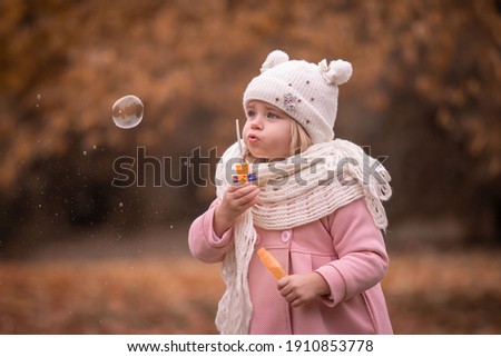 Beautiful little blonde girl, has pretty face, happy emotions, dressed in pink coat. Plays soap bubble blower. Child portrait. Lifestyle instagram concept. Autumn time. Fashion kid style.