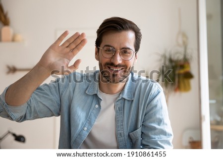Smiling millennial male in glasses posing before digital webcam waving hand chatting with friend using pc. Headshot portrait of active young man video blogger looking at camera broadcasting from home Royalty-Free Stock Photo #1910861455