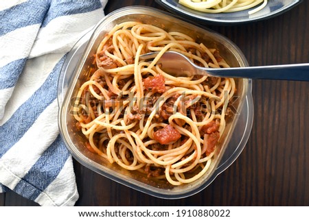 Spaghetti Bolognese with minced beef, onion, chopped tomato, garlic, olive oil, stock cube, tomato puree and Italian herb in a plastic box. Traditional Italian food in white plate with a fork.