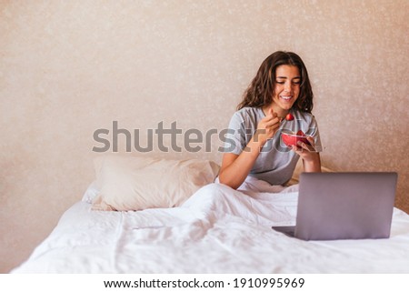 Stock photo of happy girl in pajamas using computer in the bed and eating fruit.