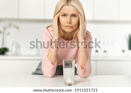 Effervescent pain reliever tablet dissolves in glass of water, a middle-aged woman in out of focus suffering from headache, holding head, massages the temples and feels hurt. Disease treatment concept